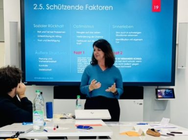 Frau in blauer Bluse erklärt Folien vor Zuhörern in einem modernen Besprechungsraum.