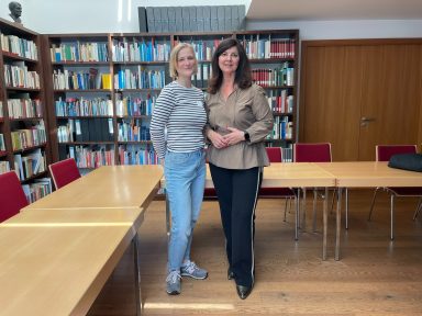 Zwei Frauen stehen in einem Bibliotheksraum vor einem Bücherregal.